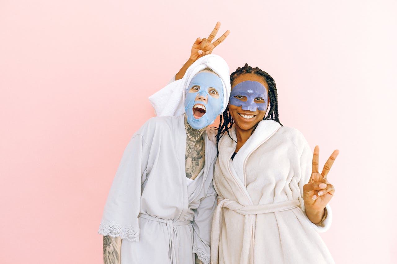 Cheerful young diverse ladies in robes and cosmetic masks applied on face standing on pale pink background and showing peace gesture while looking at camera